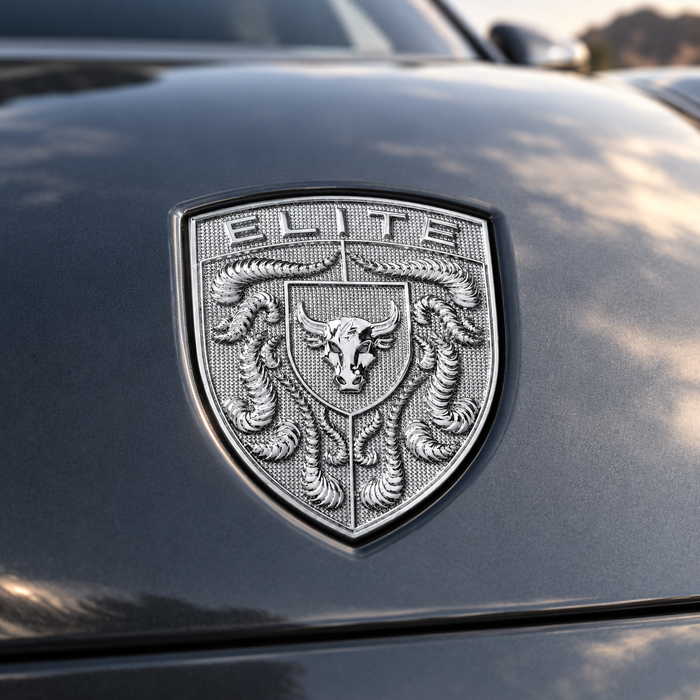 A close-up of a metallic silver shield emblem on the front hood of a dark gray vehicle. The emblem features the word 'ELITE' at the top, a central bull head, and crumrine designs.