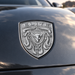 A close-up of a metallic silver shield emblem on the front hood of a dark gray vehicle. The emblem features the word 'ELITE' at the top, a central bull head, and crumrine designs.