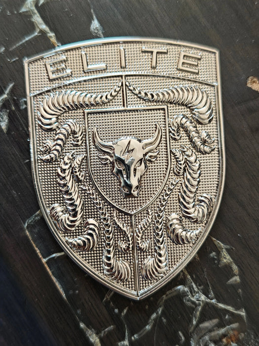 A silver metallic shield-shaped porsche badge featuring the word 'ELITE' at the top, a central bull head with a lightning bolt, and crumrine designs, photographed on a dark wood surface.