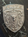 A silver metallic shield-shaped porsche badge featuring the word 'ELITE' at the top, a central bull head with a lightning bolt, and crumrine designs, photographed on a dark wood surface.