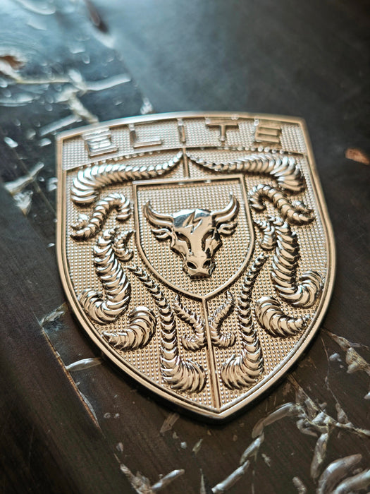 A metallic bronze shield-shaped porsche badge featuring a central bull head crest and the word 'ELITE' at the top, resting on a dark textured surface.