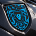 A close-up of a glossy black Porsche Mexico Blue Brahma Bull Hood Emblem featuring a bright blue shield design with a bull head, crumrine designs, and the word "ELITE" at the top, mounted on a dark metallic vehicle surface.