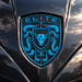 A close-up of a metallic shield-shaped emblem on a glossy black car body, featuring the word "ELITE" at the top and a stylized bull head surrounded by crumrine designs in bright blue against a black textured background.