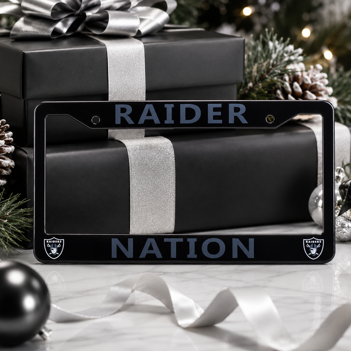 A black license plate frame with 'RAIDER NATION' in dark blue lettering and small Raiders logos in the corners, shown in a holiday gift setting with wrapped presents and pinecones.