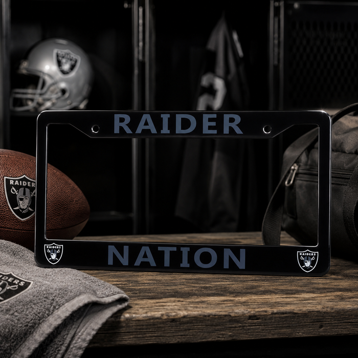 A black license plate frame with 'RAIDER NATION' in gray lettering and Oakland Raiders logos, displayed on a wooden surface with football, towel, helmet, and bag in a locker-room setting.