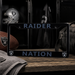 A black license plate frame with 'RAIDER NATION' in gray lettering and Oakland Raiders logos, displayed on a wooden surface with football, towel, helmet, and bag in a locker-room setting.
