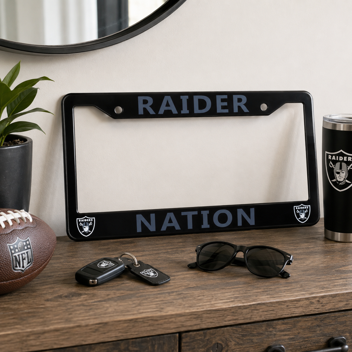 A black license plate frame with 'RAIDER NATION' in blue-gray lettering and Raiders logos, displayed on a wooden dresser alongside a football, keys, sunglasses, and a tumbler in a styled room setting.