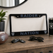 A black license plate frame with 'RAIDER NATION' in blue-gray lettering and Raiders logos, displayed on a wooden dresser alongside a football, keys, sunglasses, and a tumbler in a styled room setting.