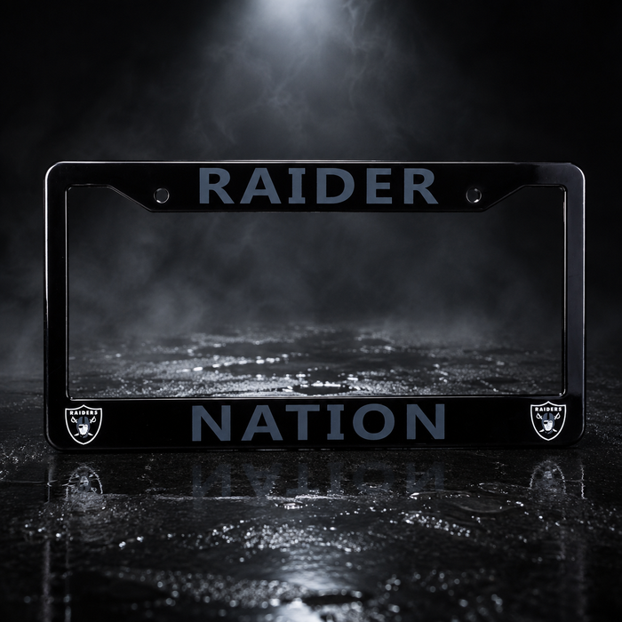 A black license plate frame featuring the words 'RAIDER NATION' in a muted blue-gray font, with the Oakland Raiders shield logos in the bottom corners. The frame is photographed on a dark, wet reflective surface with dramatic spotlighting and mist in the background.