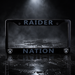 A black license plate frame featuring the words 'RAIDER NATION' in a muted blue-gray font, with the Oakland Raiders shield logos in the bottom corners. The frame is photographed on a dark, wet reflective surface with dramatic spotlighting and mist in the background.