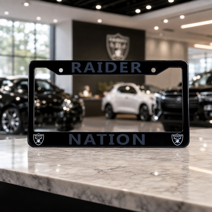 A black license plate frame with 'RAIDER NATION' and Raiders logos, displayed on a marble counter inside a car dealership showroom.