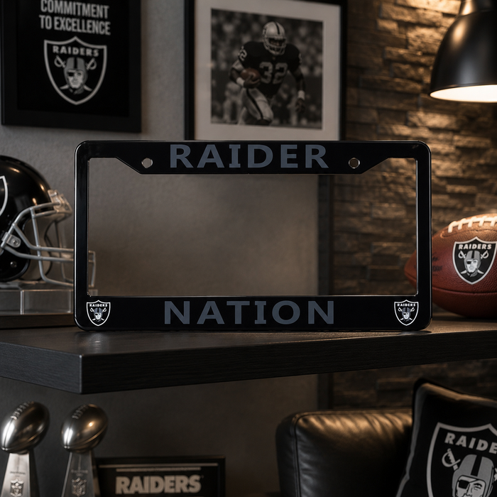 A black Raiders license plate frame with 'RAIDER' at the top and 'NATION' at the bottom, displayed on a shelf in a themed room with Raiders memorabilia.