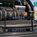 A black license plate frame with 'RAIDER NATION' and Oakland Raiders logos, displayed in the trunk of a car alongside a Raiders duffel bag, football, and tumbler.