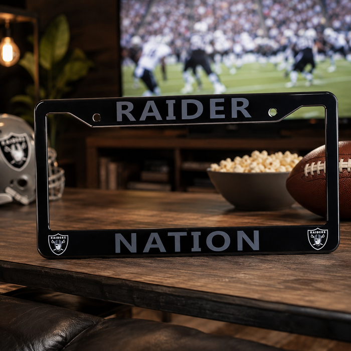 A black license plate frame featuring the words 'RAIDER NATION' in silver-gray lettering and the Oakland Raiders shield logos in the corners, displayed on a wooden table in a cozy living room setting.
