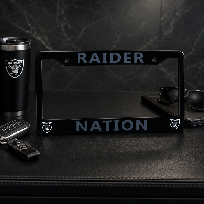 A black license plate frame with 'RAIDER NATION' and Oakland Raiders logos, displayed on a black leather surface with a matching black Raiders tumbler, keys, and sunglasses nearby.