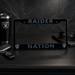 A black license plate frame with 'RAIDER NATION' and Oakland Raiders logos, displayed on a black leather surface with a matching black Raiders tumbler, keys, and sunglasses nearby.