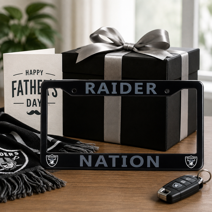 A black Raiders-themed license plate frame with 'RAIDER NATION' and team logos, displayed on a wooden table with a gift box, scarf, key fob, and Father's Day card.