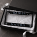 A black license plate frame with 'RAIDER NATION' and Oakland Raiders logos, presented inside an open black gift box with white tissue paper and a silver ribbon on a dark marble surface.
