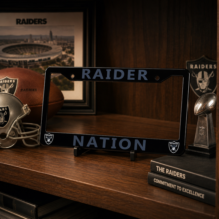 A black license plate frame featuring the words 'RAIDER' at the top and 'NATION' at the bottom, displayed on a wooden shelf alongside Raiders-themed decor including a football, helmet, framed stadium print, and trophy.