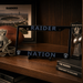 A black license plate frame featuring the words 'RAIDER' at the top and 'NATION' at the bottom, displayed on a wooden shelf alongside Raiders-themed decor including a football, helmet, framed stadium print, and trophy.