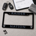 Black Raiders Nation license plate frame with silver-gray lettering and small Raiders shield logos in the corners, shown on a light gray surface with a key fob, registration document, towel, and mounting screws nearby.