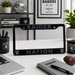 A black license plate frame with 'RAIDER NATION' printed in gray lettering and Raiders logos, displayed on a white desk in a styled office setting with Raiders-branded mugs, notebook, and decor.