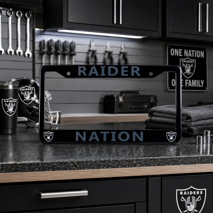 A black license plate frame with light blue Raiders branding and the words 'RAIDER NATION' is displayed on a dark kitchen counter. The background shows a matching Raiders-themed kitchen with a helmet, towel, and sign.