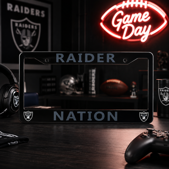 A black license plate frame with 'RAIDER' at the top and 'NATION' at the bottom, featuring the Oakland Raiders shield logo in each corner. The frame is displayed on a dark wooden desk with gaming accessories, a blurred Raiders helmet in the background, and a glowing red neon 'Game Day' sign on the wall.