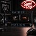 A black license plate frame with 'RAIDER' at the top and 'NATION' at the bottom, featuring the Oakland Raiders shield logo in each corner. The frame is displayed on a dark wooden desk with gaming accessories, a blurred Raiders helmet in the background, and a glowing red neon 'Game Day' sign on the wall.
