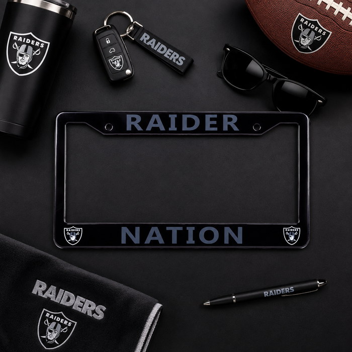 A black Raiders-themed license plate frame with 'RAIDER NATION' text and team logos, shown in a styled flat-lay with a matching tumbler, keychain, sunglasses, football, towel, and pen on a dark background.