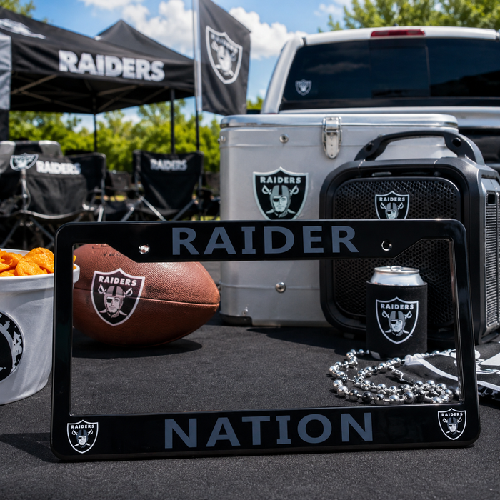 A black Raiders Nation license plate frame with silver lettering and Raiders logos, displayed in a outdoor tailgate setup with a football, cooler, speaker, and can of beer.