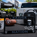 A black Raiders Nation license plate frame with silver lettering and Raiders logos, displayed in a outdoor tailgate setup with a football, cooler, speaker, and can of beer.