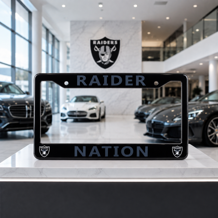 A black license plate frame with 'RAIDER NATION' in dark blue-gray lettering and small Raiders logos at the bottom corners, displayed on a showroom counter with cars and a Raiders wall sign in the background.