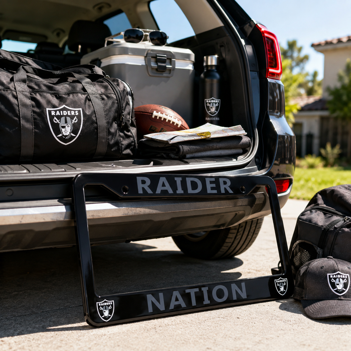 A black metal license plate frame with 'RAIDER' on top and 'NATION' on the bottom, featuring Raiders logos, displayed in front of an open SUV trunk with team gear including a bag, cooler, water bottle, football, and a cap.