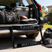A black metal license plate frame with 'RAIDER' on top and 'NATION' on the bottom, featuring Raiders logos, displayed in front of an open SUV trunk with team gear including a bag, cooler, water bottle, football, and a cap.