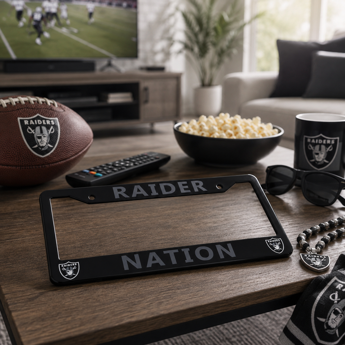 A black metal license plate frame engraved with 'RAIDER NATION' and featuring Oakland Raiders logos, displayed on a wooden coffee table in a living room setting alongside a Raiders football, popcorn, sunglasses, and a matching mug.
