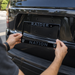 A person's hands are shown installing a black license plate frame onto the rear of a black SUV. The frame features the word 'RAIDER' at the top and 'NATION' at the bottom, with the Oakland Raiders shield logo on each corner.
