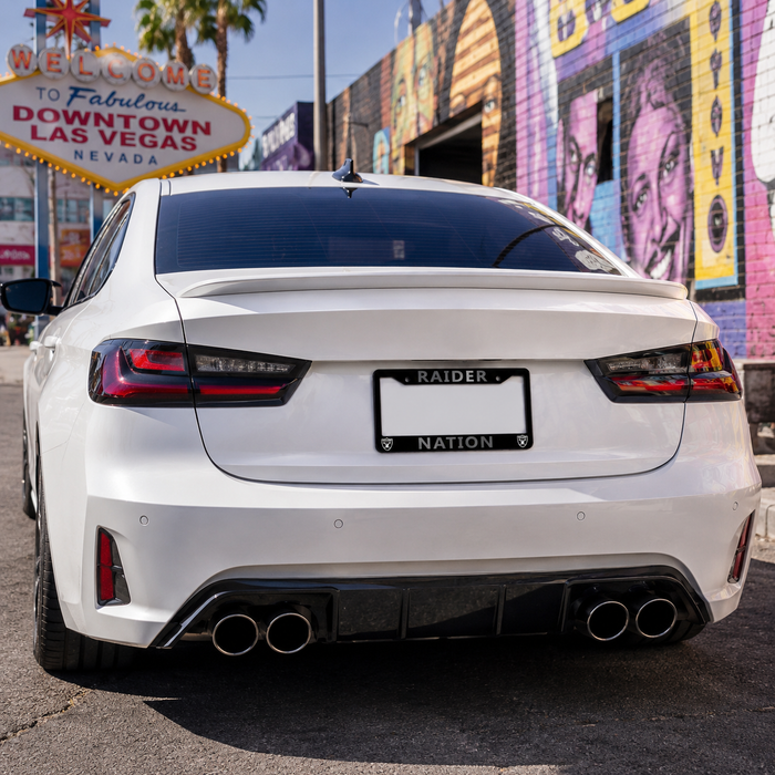 A white sedan shown from the rear in an outdoor street setting, with a black license plate frame reading 'RAIDER NATION' mounted behind the plate. The car has a rear spoiler, dual exhaust pipes, and LED taillights.