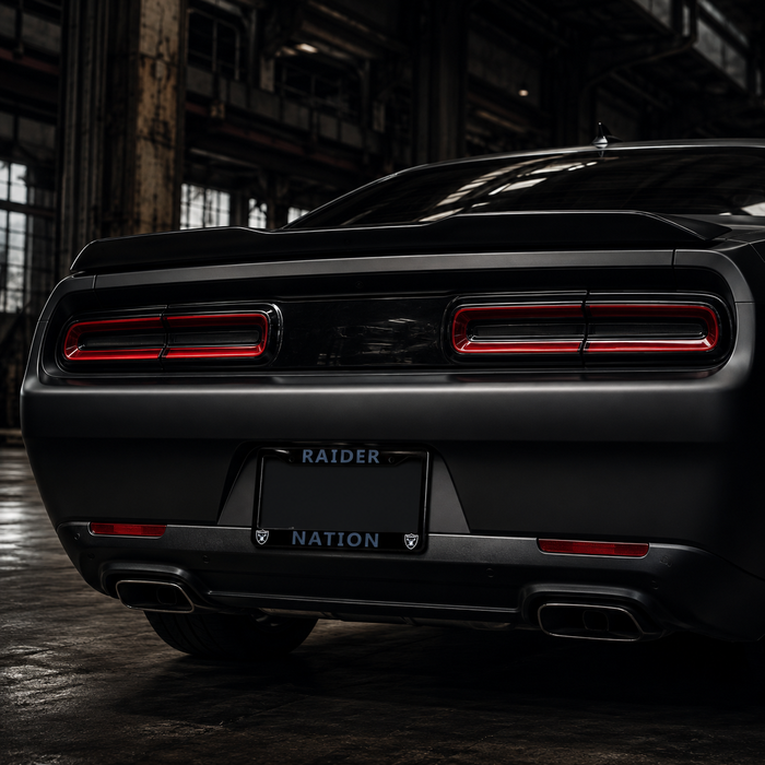 Rear view of a matte black muscle car in a dark industrial warehouse setting, featuring horizontal taillights, a black license plate frame reading 'RAIDER NATION', and dual exhausts.