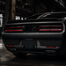 Rear view of a matte black muscle car in a dark industrial warehouse setting, featuring horizontal taillights, a black license plate frame reading 'RAIDER NATION', and dual exhausts.