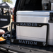 A black metal license plate frame with 'RAIDER NATION' and Oakland Raiders logos, displayed on the tailgate of a truck next to a white cooler.