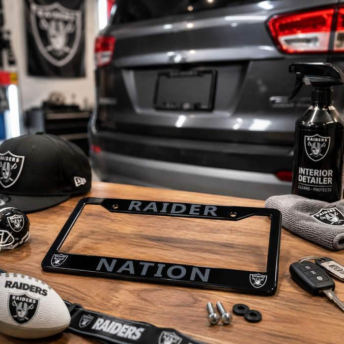 A black Raiders Nation license plate frame with silver lettering is displayed on a wooden table, surrounded by Raiders-themed accessories including a black Raiders cap, a football, a towel, and a spray bottle of interior detail cleaner. The background shows the rear of a gray vehicle in a garage setting.