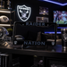 A black metal license plate frame with 'RAIDER NATION' printed in silver, displayed on a dark shelf in a themed room with Raiders memorabilia, including a helmet, football, and a matching tumbler.