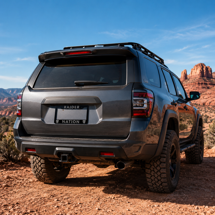 A dark gray Jeep Gladiator pickup truck with a roof rack and off-road tires, shown from the rear three-quarter view on a dirt trail with red rock desert formations in the background. The license plate reads 'RAIDER' and 'NATION'.