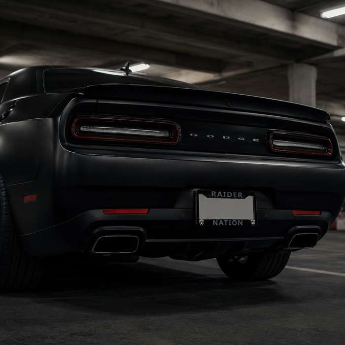 Rear view of a matte black Dodge Challenger in a parking garage, featuring the rear license plate frame with 'RAIDER NATION' text, dual exhausts, and the Dodge logo.