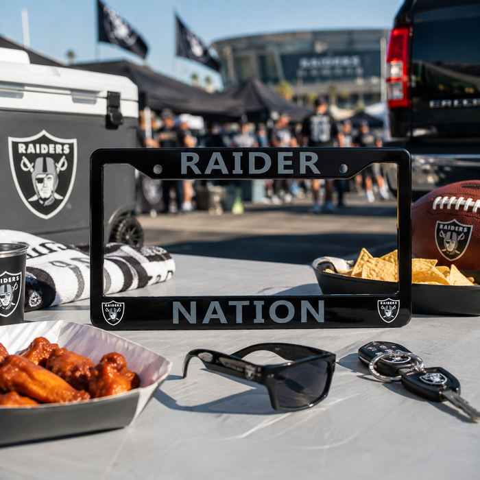 A black metal license plate frame with 'RAIDER NATION' and Oakland Raiders logos, displayed on a table with Raiders-branded accessories including a cooler, sunglasses, keys, chips, and wings in an outdoor event setting.