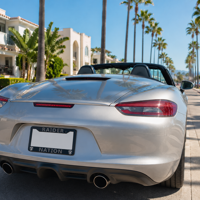 Silver Porsche Roadster Convertible shown from the rear, parked on a sunny street with palm trees and a Mediterranean-style building in the background. The license plate frame reads 'RAIDER NATION'.