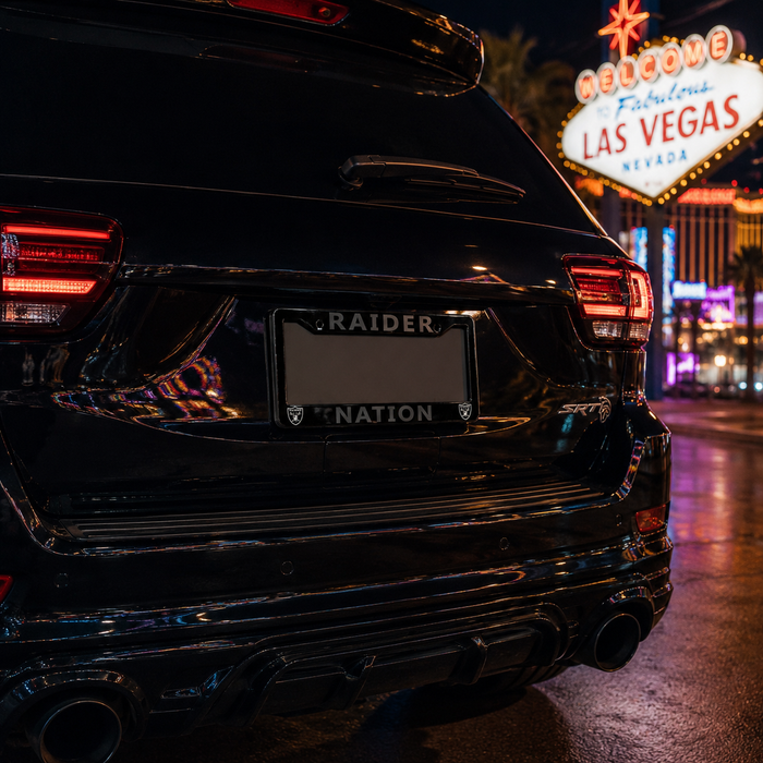 Rear view of a glossy black SUV parked on a city street at night, with the license plate frame reading "RAIDER" and "NATION" below it. The Fabulous Las Vegas sign is visible in the background.