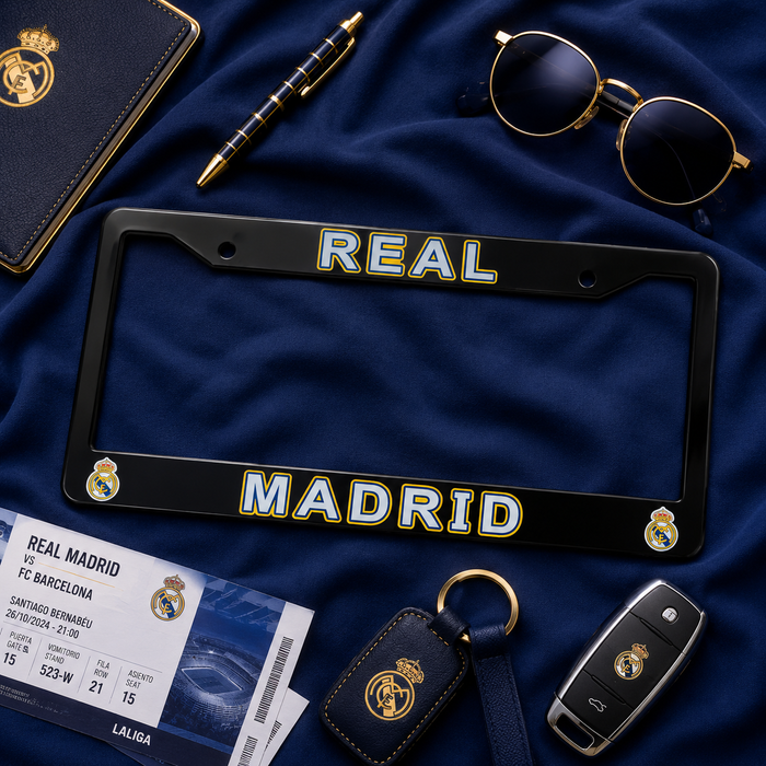A black license plate frame with 'REAL MADRID' in white and gold lettering, styled on a dark blue fabric background with Real Madrid-themed accessories including a passport page, key fob, leather key case, pen, and sunglasses.