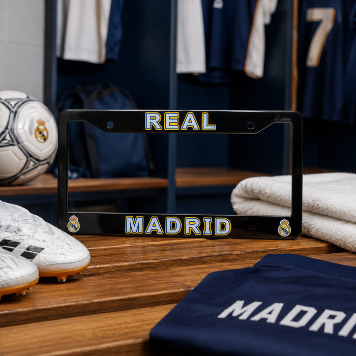A black license plate frame with 'REAL MADRID' in gold and white lettering, displayed on a wooden bench in a Real Madrid locker room setting with soccer shoes, a soccer ball, and a navy jersey nearby.